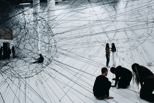 People exploring a white room filled with dense black strings forming large web‑like shapes.