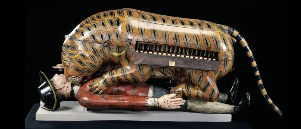 A painted wooden tiger automaton pinning a uniformed figure on his back, with visible keyboard-like mechanisms in the tiger’s side.