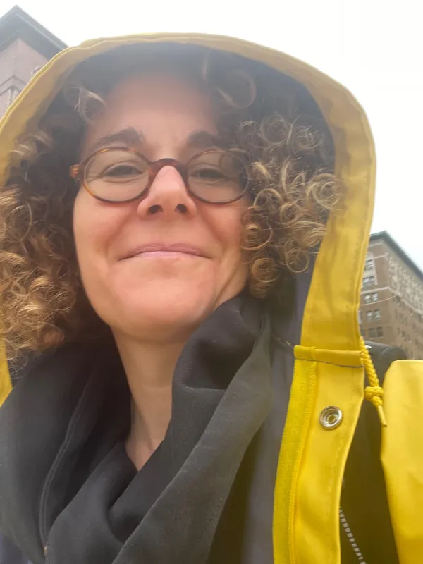 A woman weraing a yellow raincoat, with curly hear and glasses, is smiling into the camera
