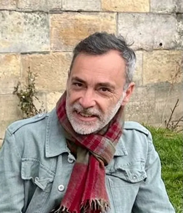a bearded man, wearing a scarf and a jacket, in front of a brick wall