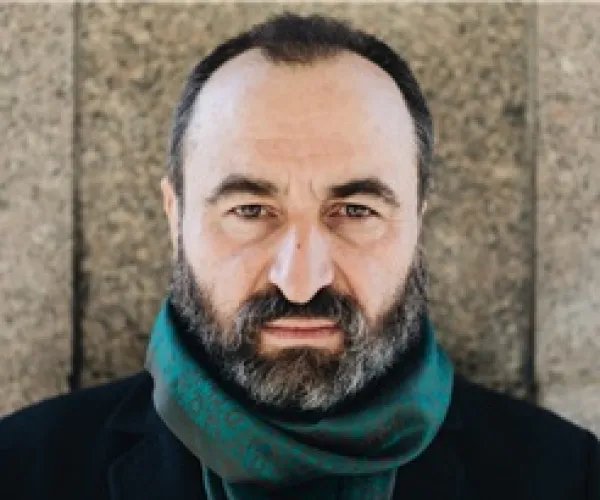 the photo shows a bearded man, looking solemnly into the camera. He wears a greenish scarf