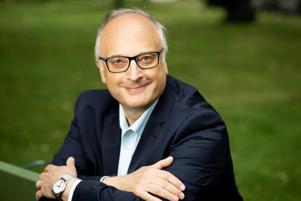 Franco De Angelis smiling while sitting arms crossed, wearing eyeglasses and a blue suit.