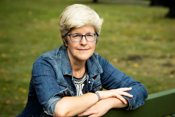 Helen Beebee seated on a park bench, wearing eyeglasses and a blue denim jacket.