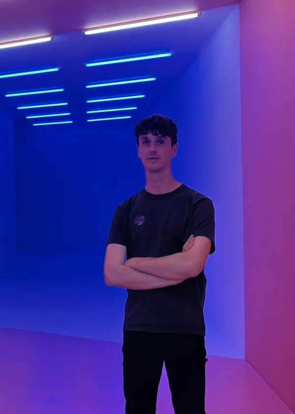 Mark standing in a dimly lit room with blue walls. His arms are crossed.