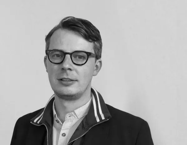 Jason Waite in a collared shirt and jacket with striped collar detail, standing against a plain light-colored wall in a black-and-white photo.