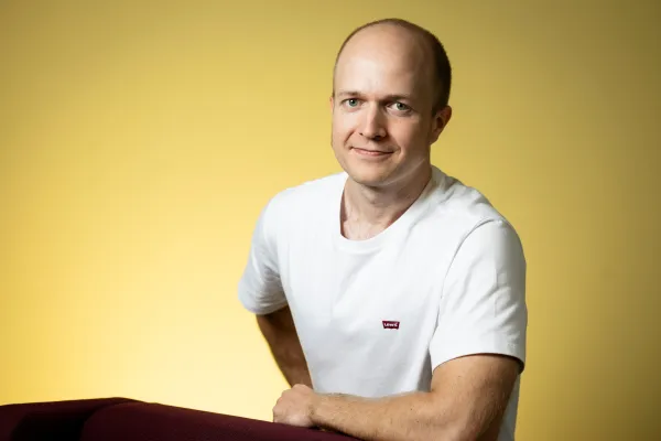 Roope Kaaronen. Photo by V. Somerpuro. Roope Kaaronen smiling with one hand on his hip and the other resting on the back of a chair.