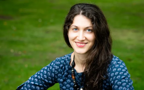 Maria Khachaturyan, wearing a blue shirt and a wooden necklace, smiling widely.