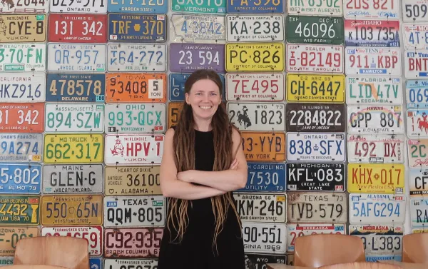 Ksenia Medvedeva. Ksenia Medvedeva standing in front of a wall made of U.S. license plates, arms crossed and smiling broadly.
