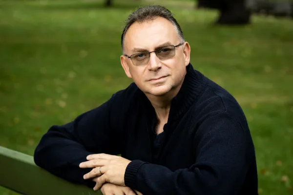 Paul Froshseated on a park bench, arms resting on the backrest, wearing glasses and a black button-up shirt.