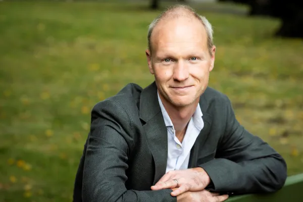Tim Stuart-Buttle. Photo by V. Somerpuro. Tim Stuart-Buttle in a dark blazer and white shirt, leaning on a green bench.