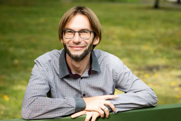 Patryk Dziurosz-Serafinowicz. Photo by V. Somerpuro. Patryk Dziurosz-Serafinowicz, wearing glasses and a button-up shirt, sits on a green bench and smiles.