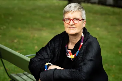 Friederike Lüpke, wearing red eyeglasses and a colorful necklace, sitting on a green park bench.