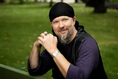 Frog sitting on a park bench, wearing a black headscarf, dark leather vest, and jewelry.
