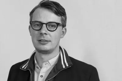 Jason Waite in a collared shirt and jacket with striped collar detail, standing against a plain light-colored wall in a black-and-white photo.