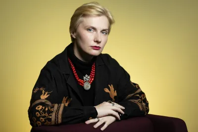 Tetiana Kalytenko, wearing a black jacket with brown patterned sleeves and a red beaded necklace with a large pendant, hands resting on the arm of a chair.