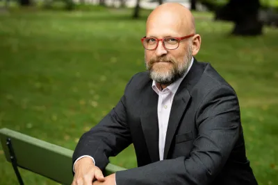 Kirby Deater-Deckard seated on a park bench, arms resting on the backrest, wearing red glasses and a gray suit.