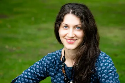 Maria Khachaturyan, wearing a blue shirt and a wooden necklace, smiling widely.