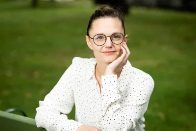 Mona Mannevuo wearing glasses and a white button-up shirt, resting her face against her hand and smiling.
