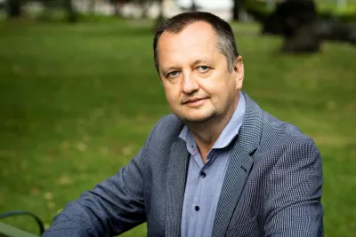 Piotr Żuk wearing a light blue shirt and patterned blazer, seated outdoors on a green bench.