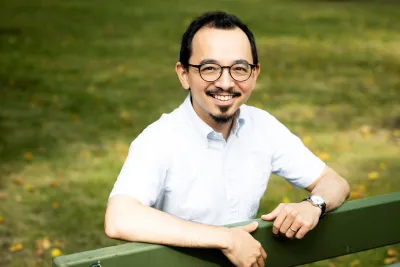 Michael Yoshitaka Erlewine, wearing a white button-up shirt and glasses, , sits on a green bench and smiles.