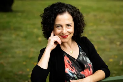 Emma Polyakov wearing a colorful shirt and red lipstick, smiling with two fingers resting on her temple.